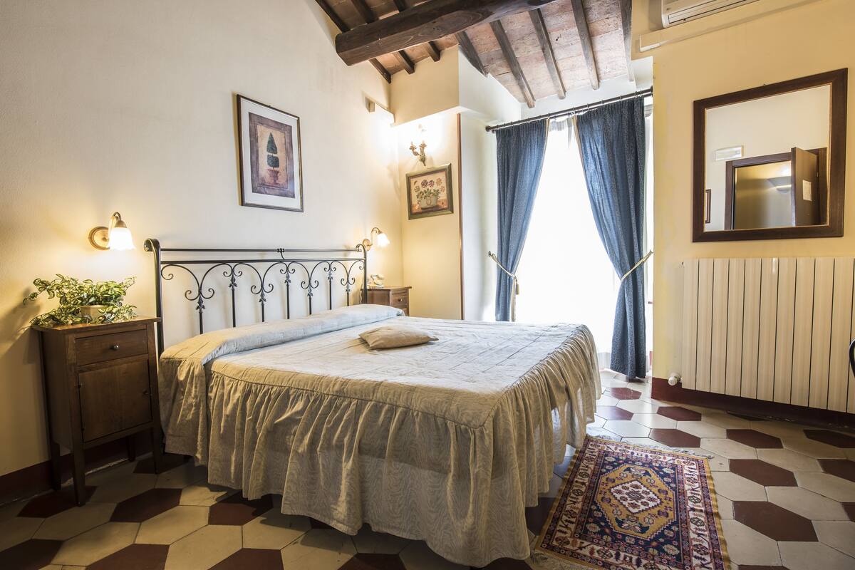 A bedroom with a bed, a nightstand, a wall-mounted mirror, and a window with curtains.