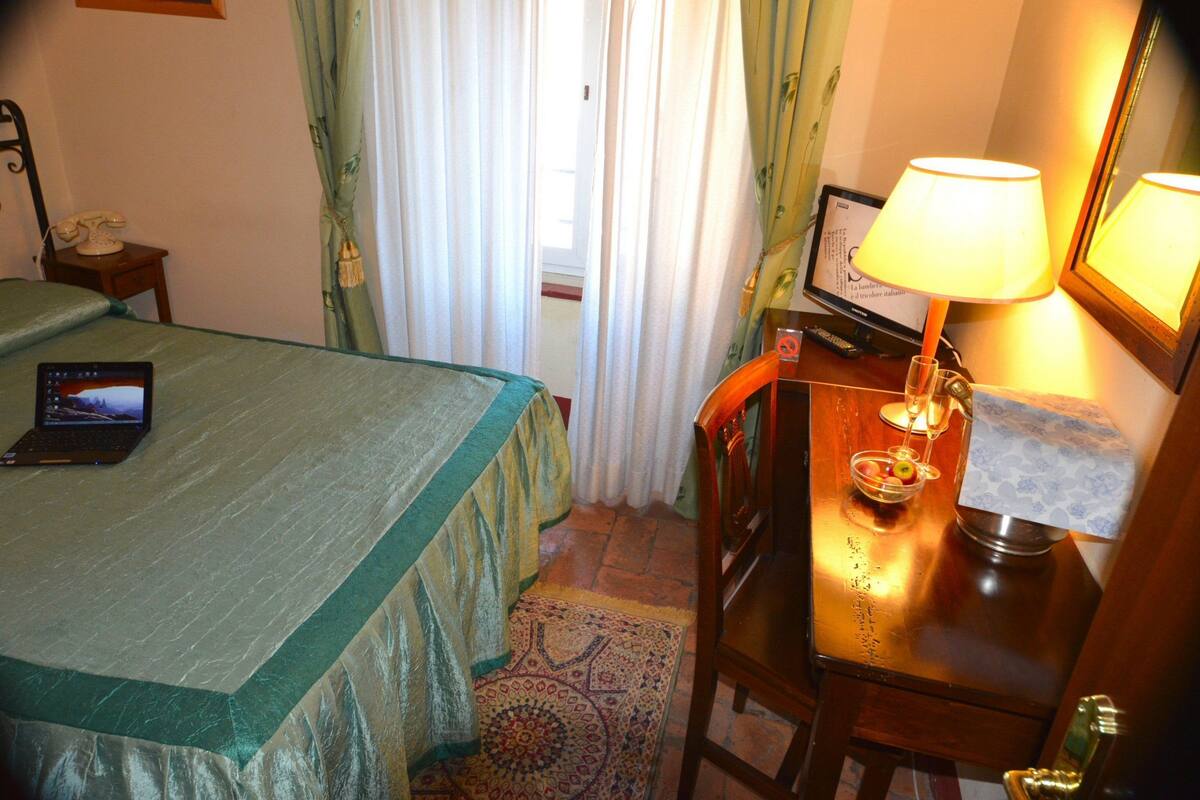 A hotel room with a bed, a desk with a laptop, a lamp, and a small table with a bowl and a glass.