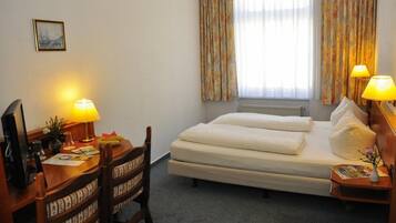 A hotel room with a bed, desk, chair, lamps, and a window with curtains.