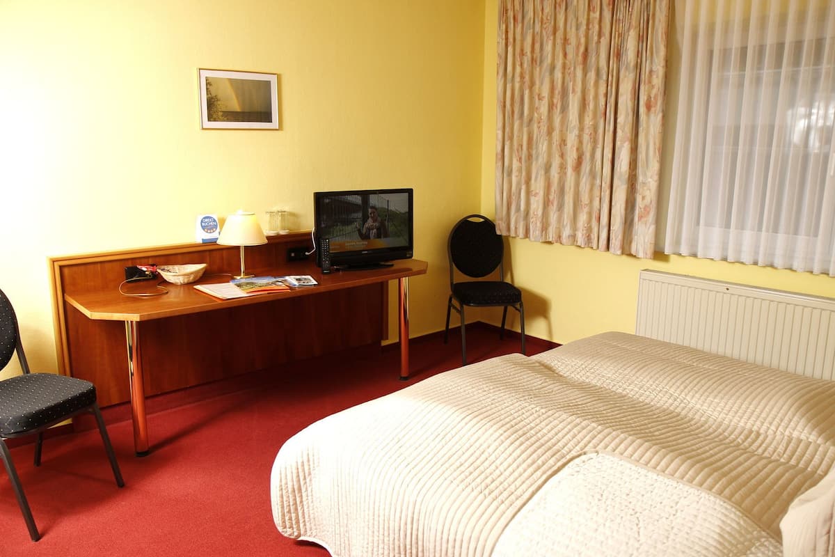 Ein Hotelzimmer mit einem Bett, einem Schreibtisch mit Fernseher, einem Sessel und einem Fenster mit Vorhängen.