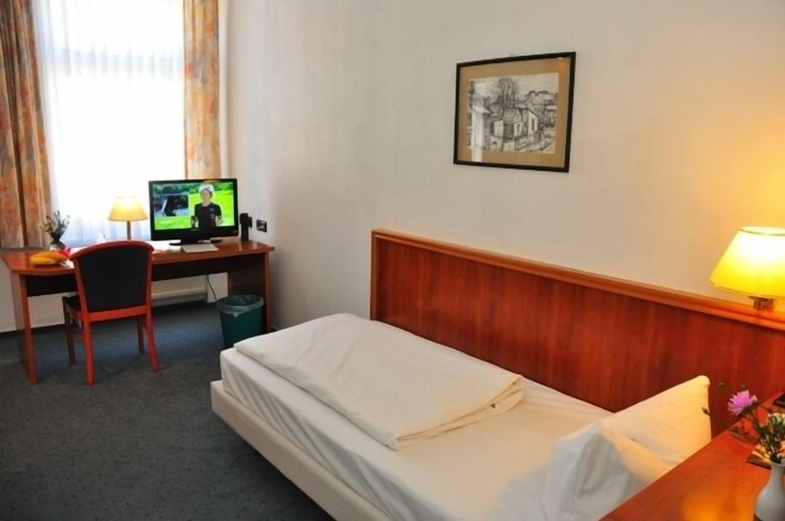 A hotel room with a bed, a desk with a computer, a chair, a lamp, and a framed picture on the wall.