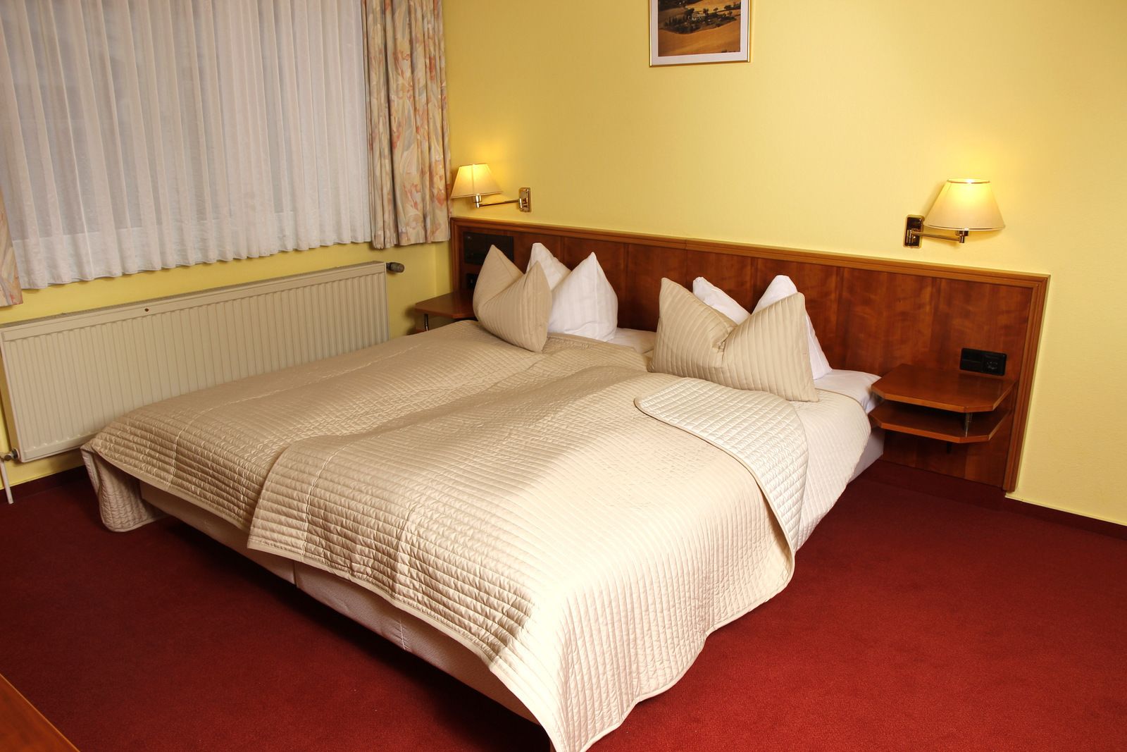 Habitación de hotel con una cama grande, dos mesitas de noche, cabecera de madera, un radiador y dos lámparas fijadas a la pared.