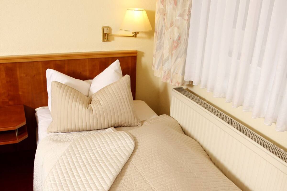 Ein ordentlich bezogenes Bett mit einer gesteppten Decke und Kissen, einer Nachttischlampe und einer Heizung.