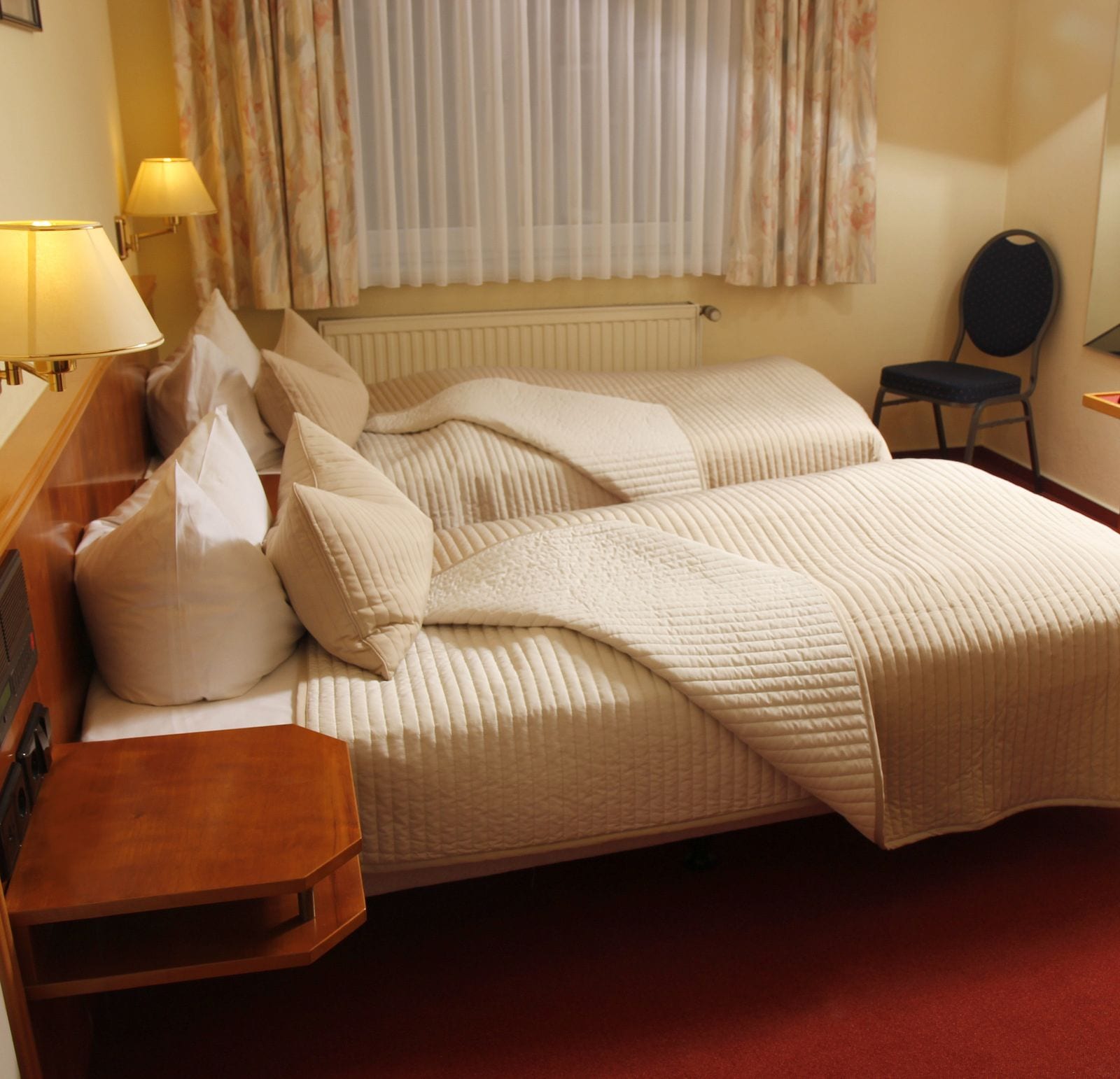 Habitación de hotel con cama, dos mesitas de noche, una silla, una lámpara y ventana con cortinas.