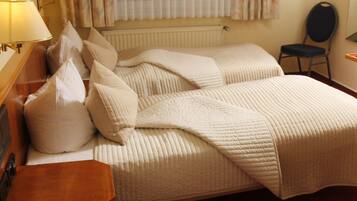 Habitación de hotel con cama, dos mesitas de noche, una silla, una lámpara y ventana con cortinas.