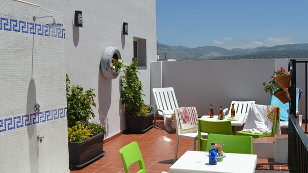 Una terraza con un muro blanco de baldosas, una fuente de agua, plantas en macetas y mobiliario exterior con mesa y sillas.