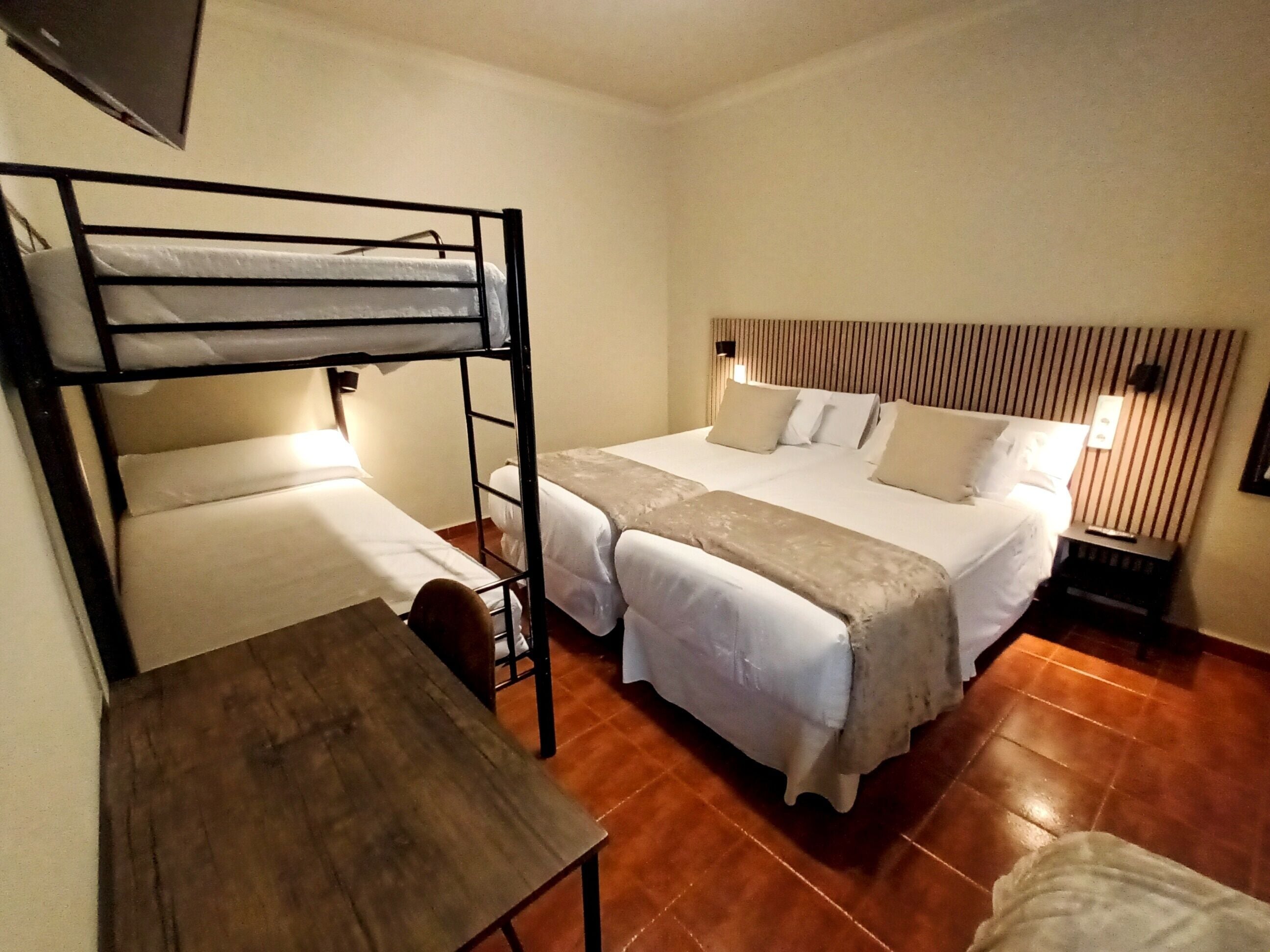 Una habitación de hotel con una cama litera, una cama individual, una mesa de madera y una mesita de noche con lámpara.