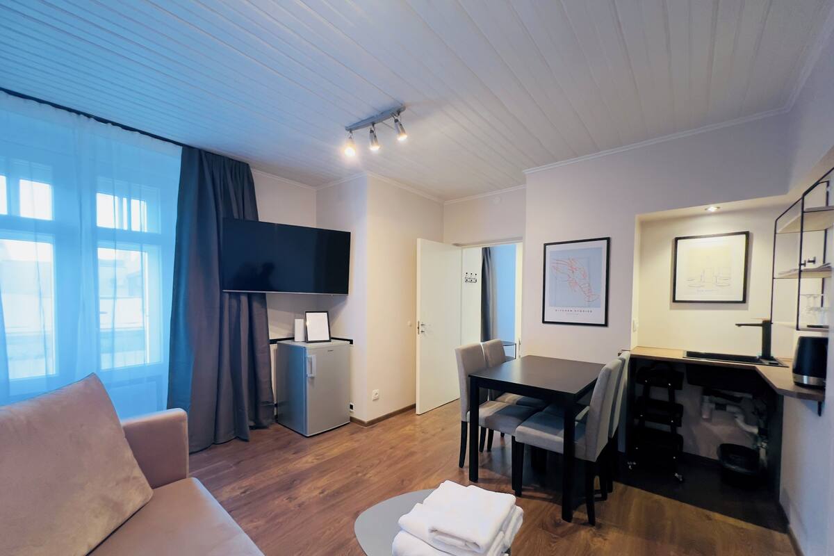 A modern hotel room with a sofa, a dining table, a small kitchen area, and a TV mounted on the wall.