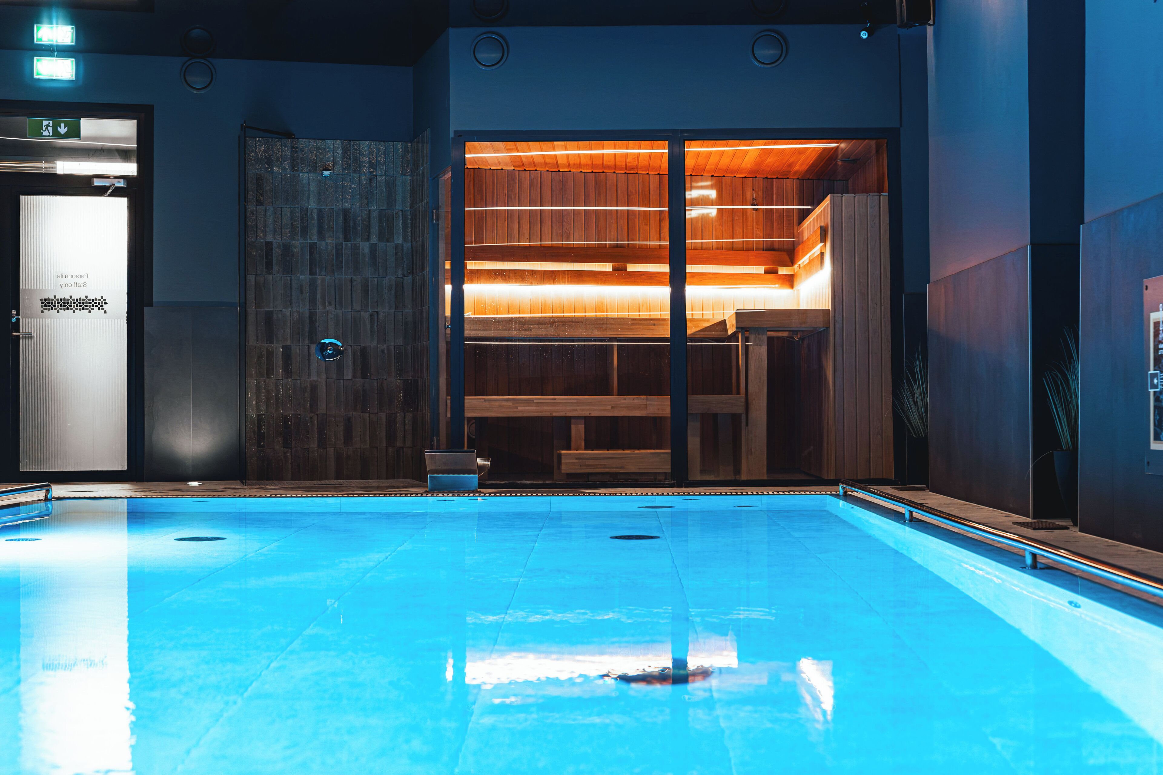 Une piscine intérieure moderne, dotée d’une eau claire, de bancs en bois et d’un espace sauna avec des murs et des bancs en bois.