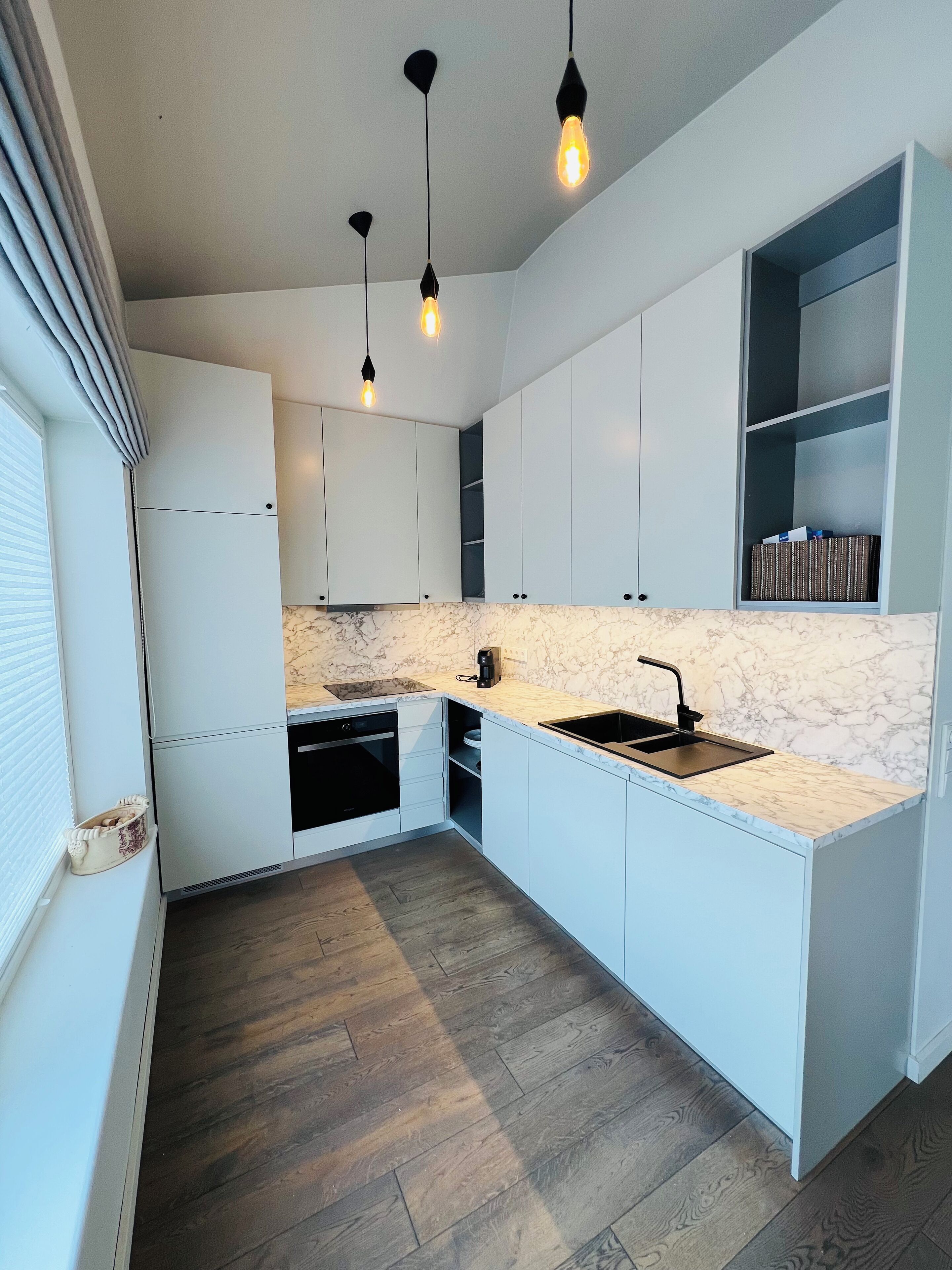 Una cocina moderna con gabinetes blancos, encimera de mármol y horno empotrado. Tres lámparas colgantes iluminan la encimera.