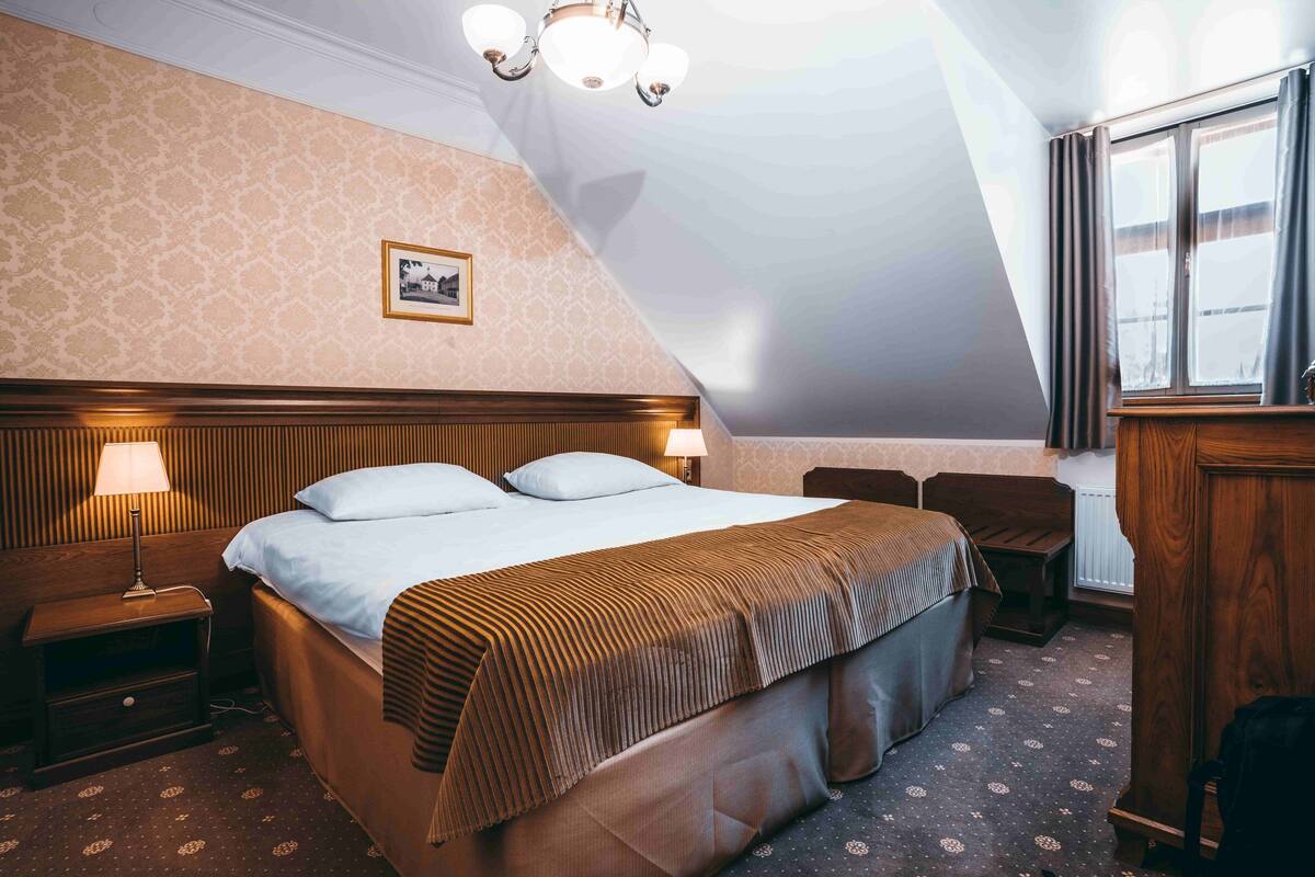 A hotel room with a large bed, two bedside tables with lamps, a wooden headboard, a picture on the wall, and a window with curtains.