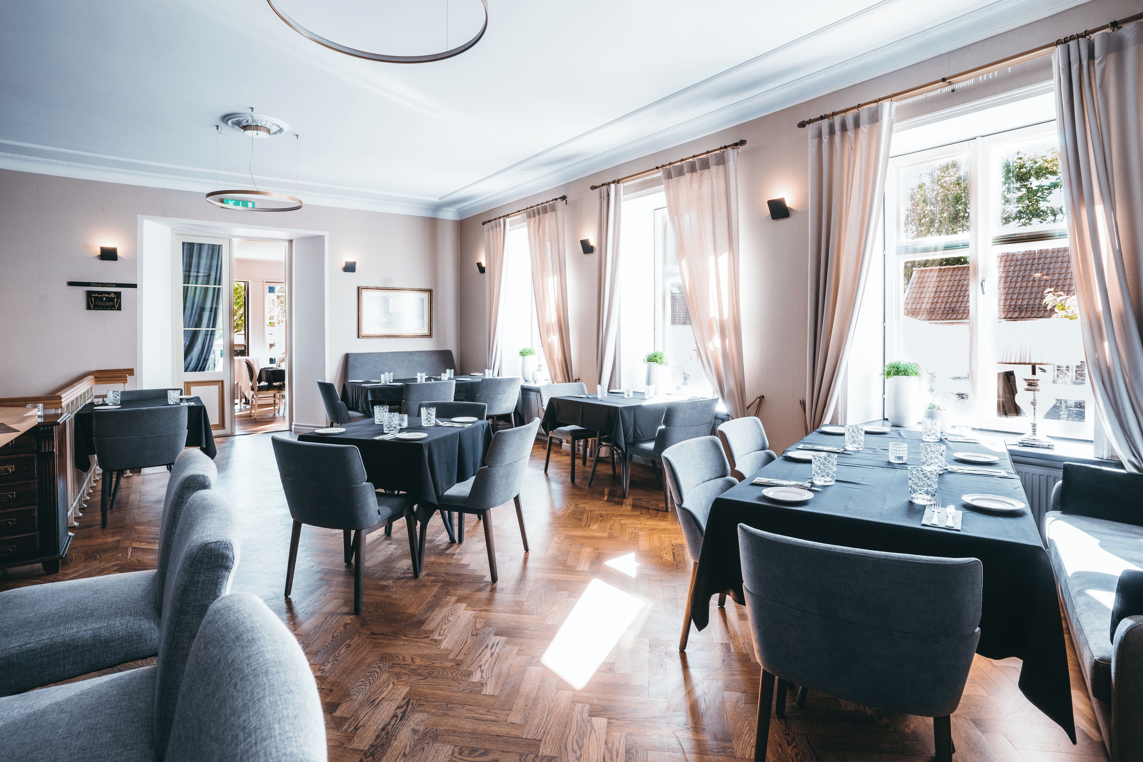 Un comedor amplio con varias mesas y sillas, ventanas grandes con cortinas, y piso de madera.