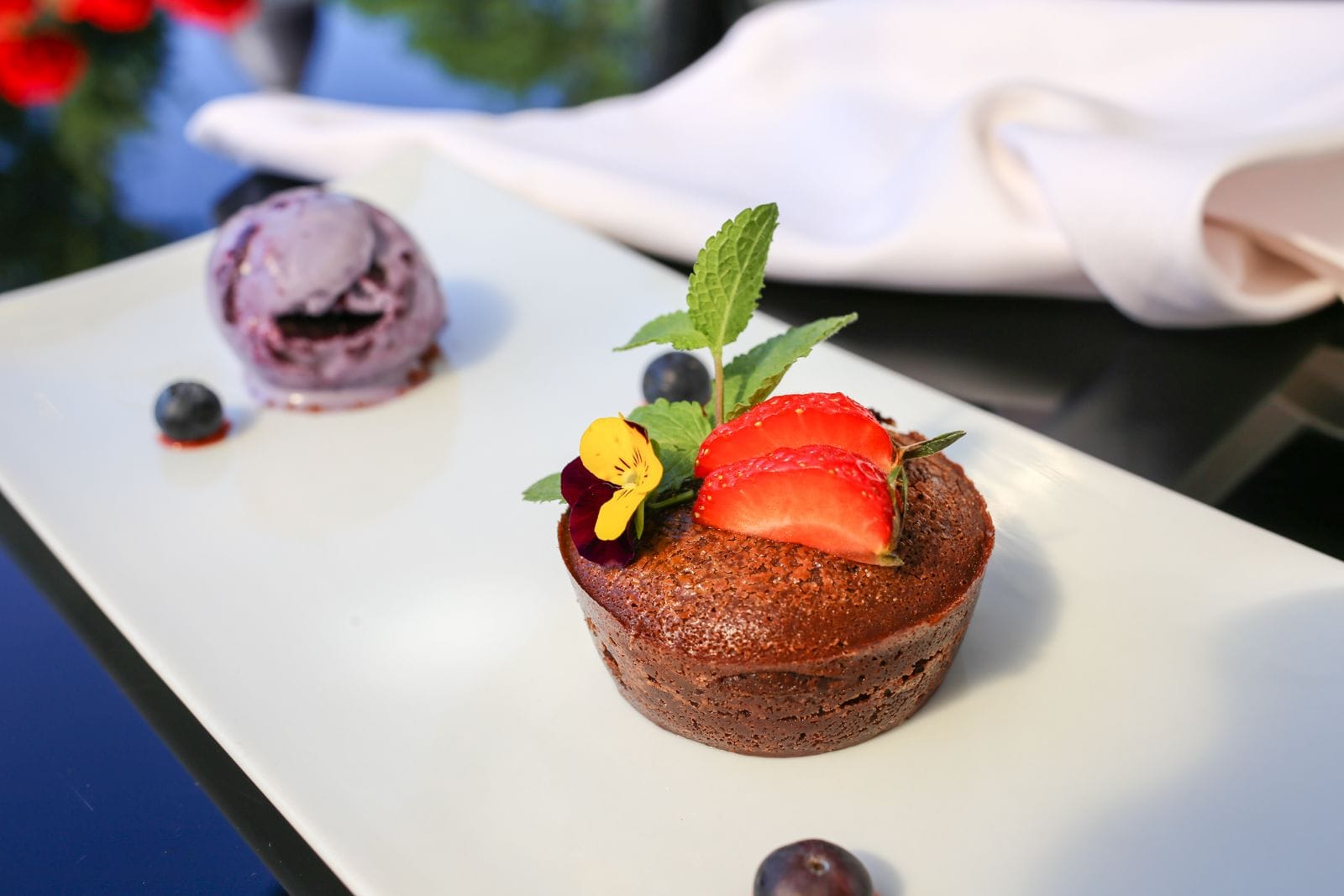 Un plato de postre con pastel de chocolate cubierto con fresa y menta, acompañado de una porción de helado de arándanos y un arándano.