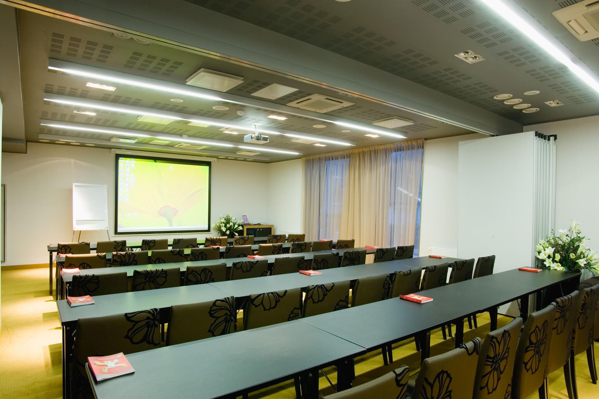 Un salón de conferencias con una larga mesa, filas de sillas, una pantalla de proyector y una pizarra.