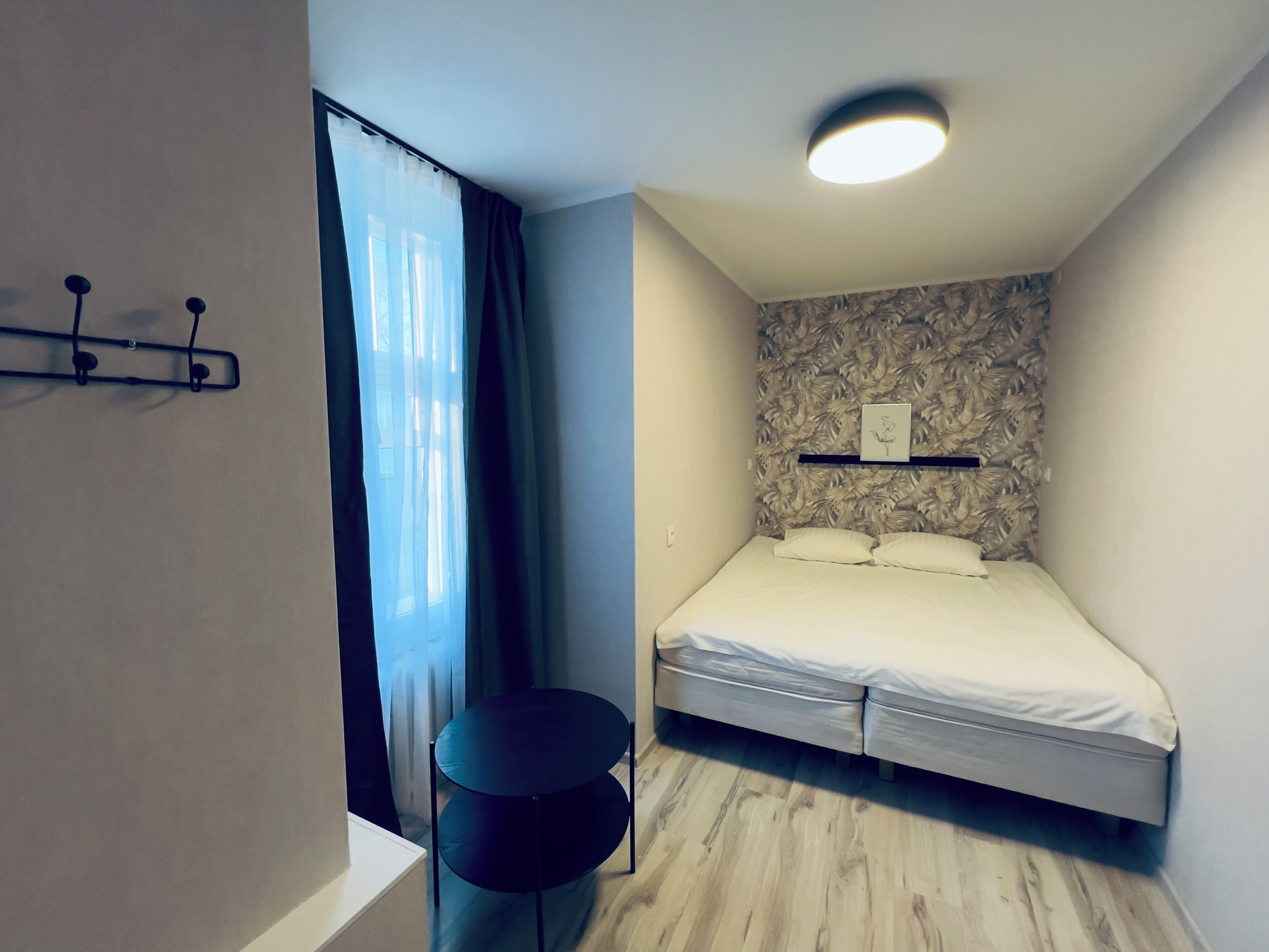 A bedroom with a bed, a small table, a wall-mounted shelf, and a ceiling light.