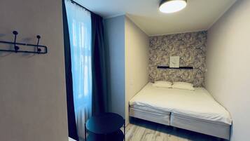 A bedroom with a bed, a small table, a wall-mounted shelf, and a ceiling light.