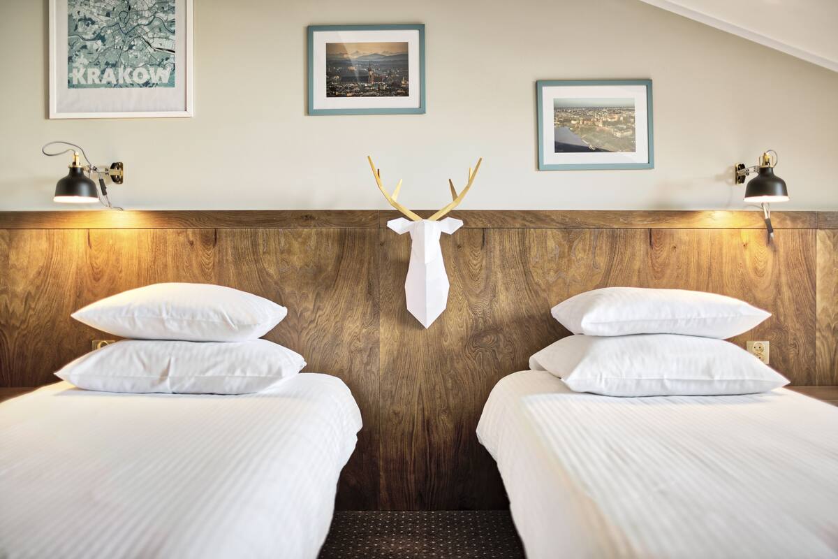 Hotelkamer met twee bedden, een houten hoofdeinde, witte beddengoed, ingelijste foto's aan de muur en een decoratie van een hertenkop.