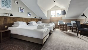 Une chambre d’hôtel avec un grand lit, deux petits lits, un bureau et une chaise. Les murs sont ornés de tableaux encadrés et une fenêtre laisse entrer la lumière naturelle.