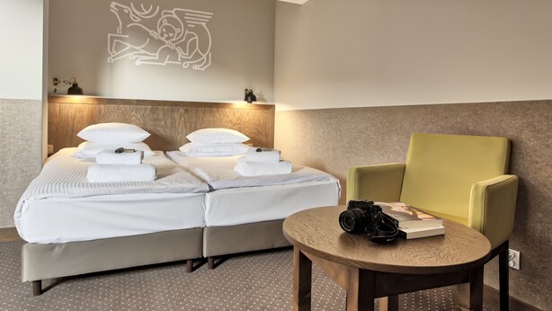 Een netjes opgemaakt bed met witte lakens en een kussen, een groene fauteuil en een kleine tafel met een camera en een boek.