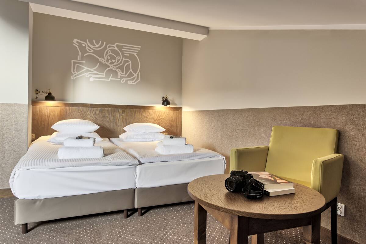 Een net opgemaakt bed met witte lakens en kussens, een houten hoofdbord, een groene fauteuil en een klein rond tafeltje met een camera en een boek.