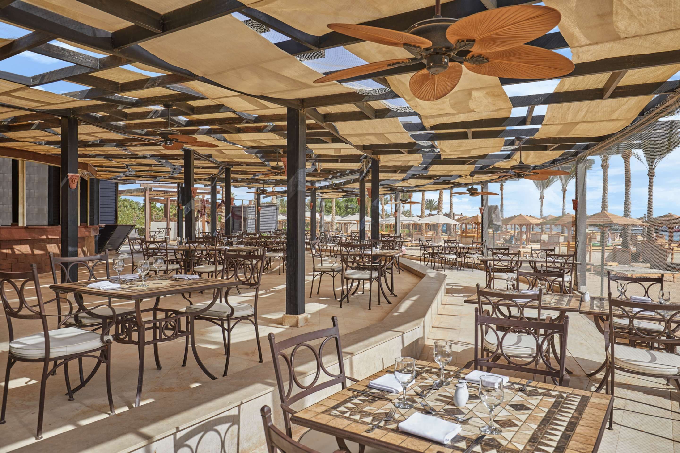 An outdoor restaurant with multiple tables and chairs, a ceiling fan, and a view of palm trees and buildings in the background.
