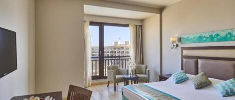 A hotel room with a bed, a desk with a fruit bowl, a chair, a small table, and a balcony with a view of buildings.