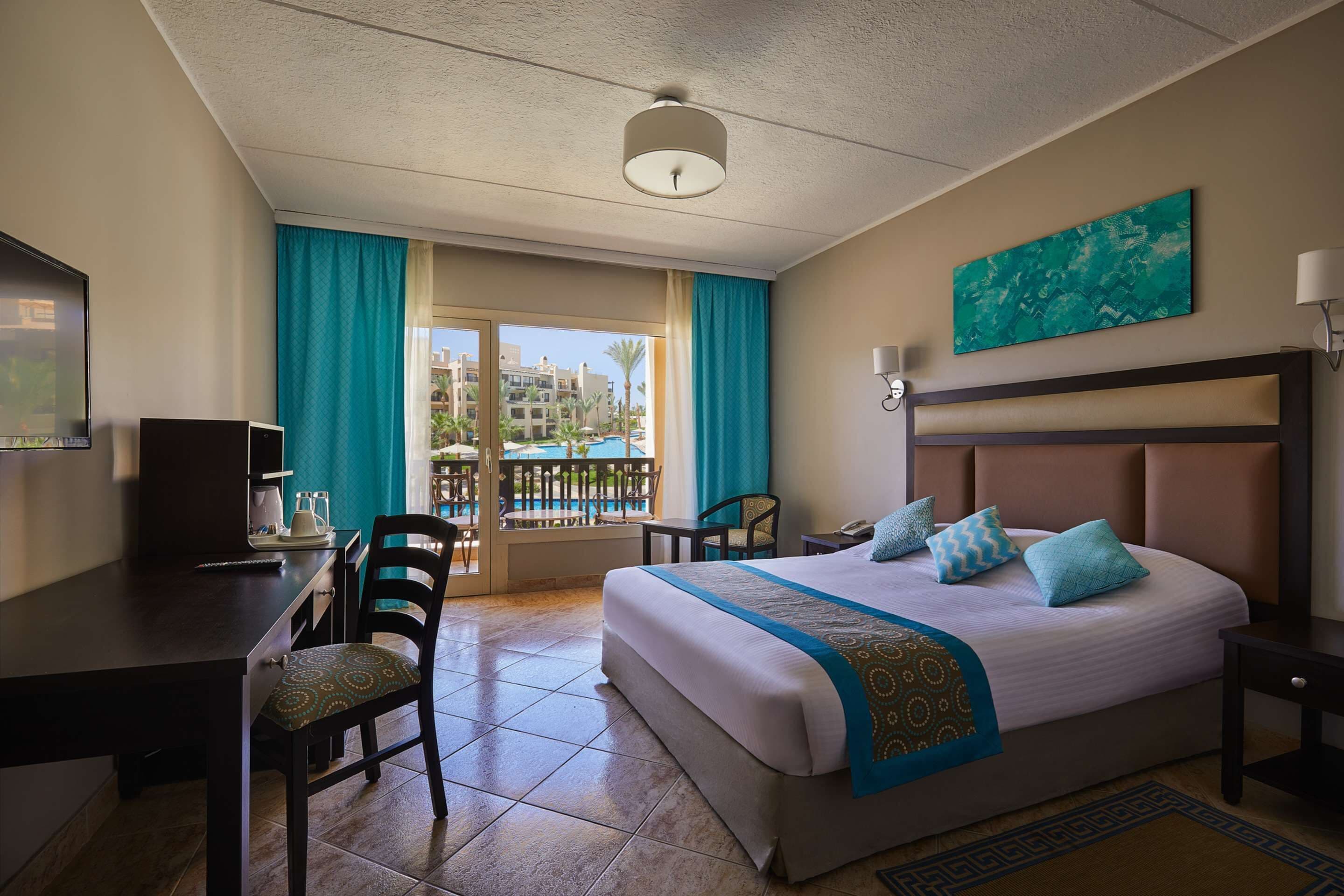 Habitación de hotel con cama, escritorio, sillas, televisor y balcón con vistas a edificios y palmeras.