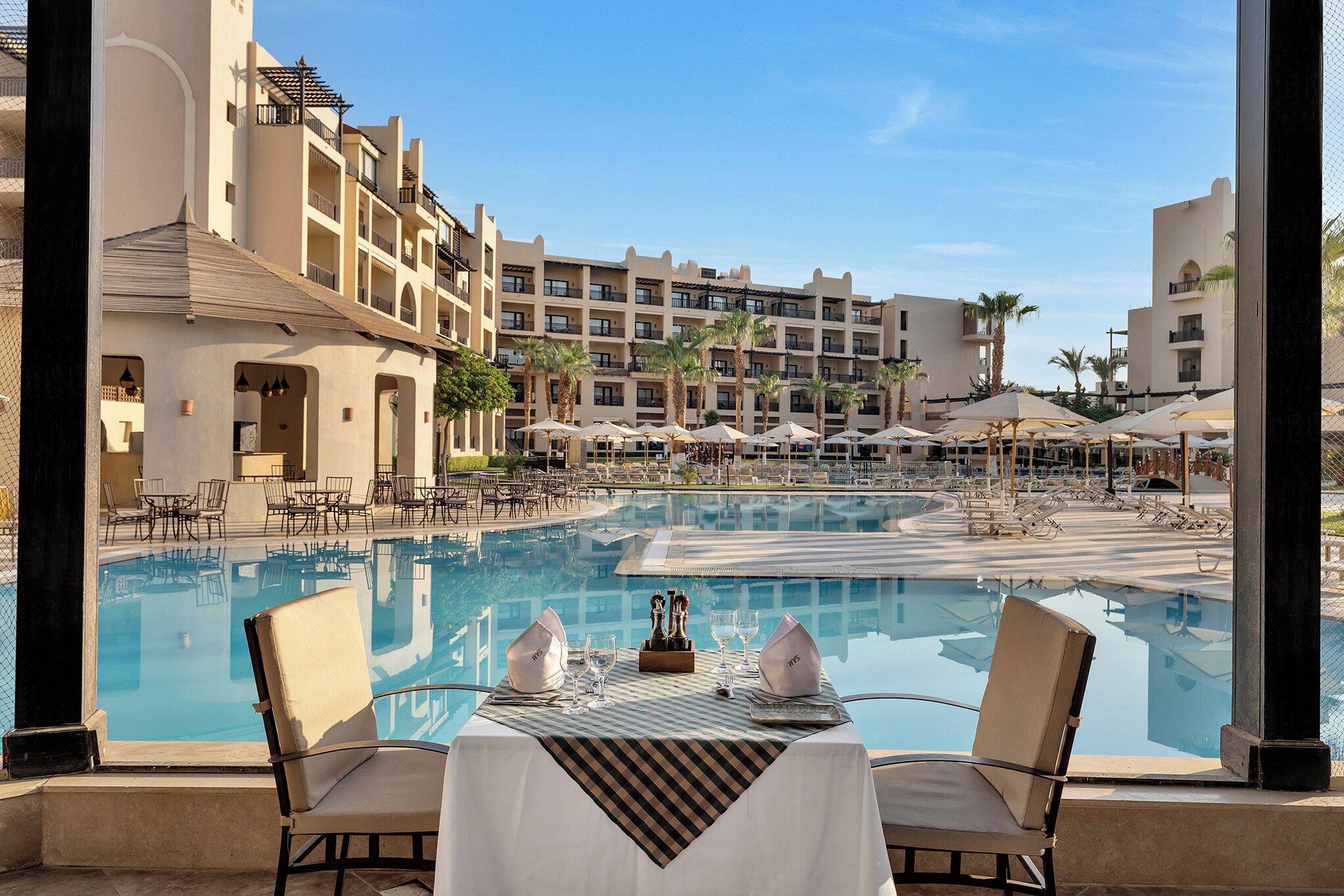 A poolside dining area with a table set for two, overlooking a large pool and a hotel building with multiple balconies.
