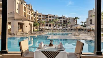 A poolside dining area with a table set for two, overlooking a large pool and a hotel building with multiple balconies.