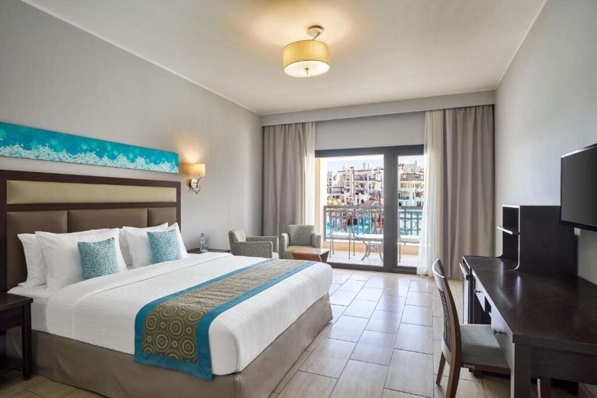 A hotel room with a bed, a desk, a chair, a TV, a balcony with a view of buildings, and a painting on the wall.