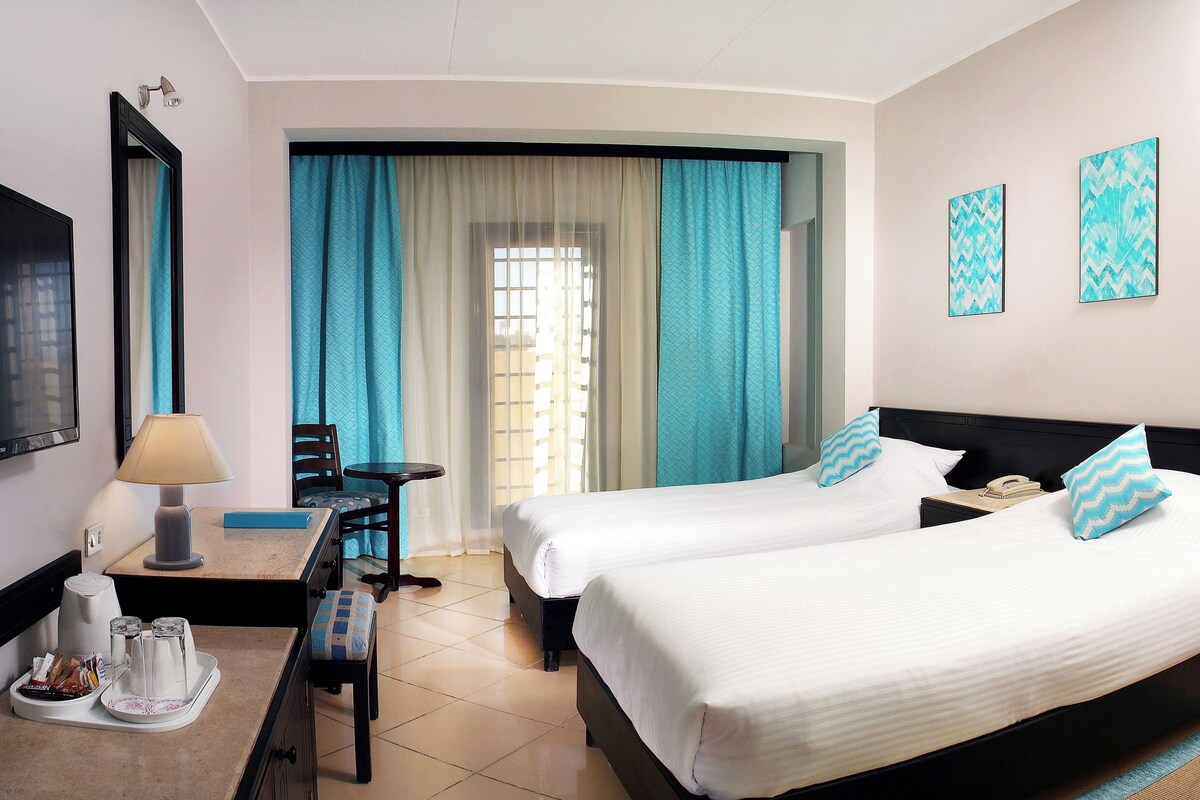 A hotel room with two beds, a desk with a lamp, a chair, a TV, and artwork on the wall.