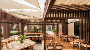 Un área de comedor al aire libre con mesas y sillas de madera, cubierta por una pérgola de madera con ventilador de techo.