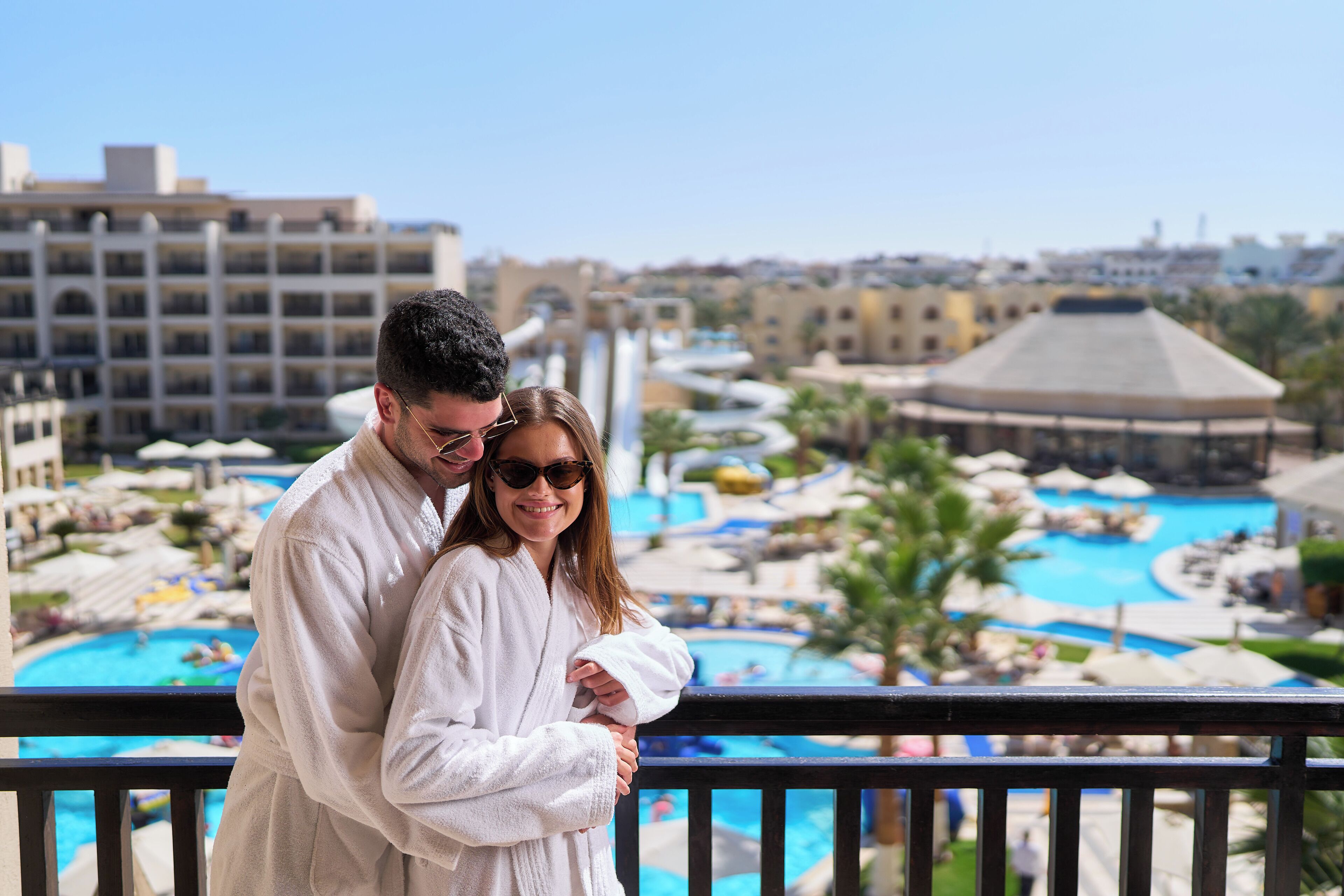 Una pareja parada en un balcón con vistas a un área de piscina de un resort, con varias piscinas y sillas de descanso.