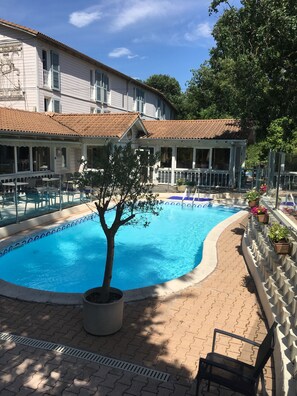 Una piscina con solárium de baldosas, un árbol en maceta y un edificio con tejado y ventanas de baldosas.