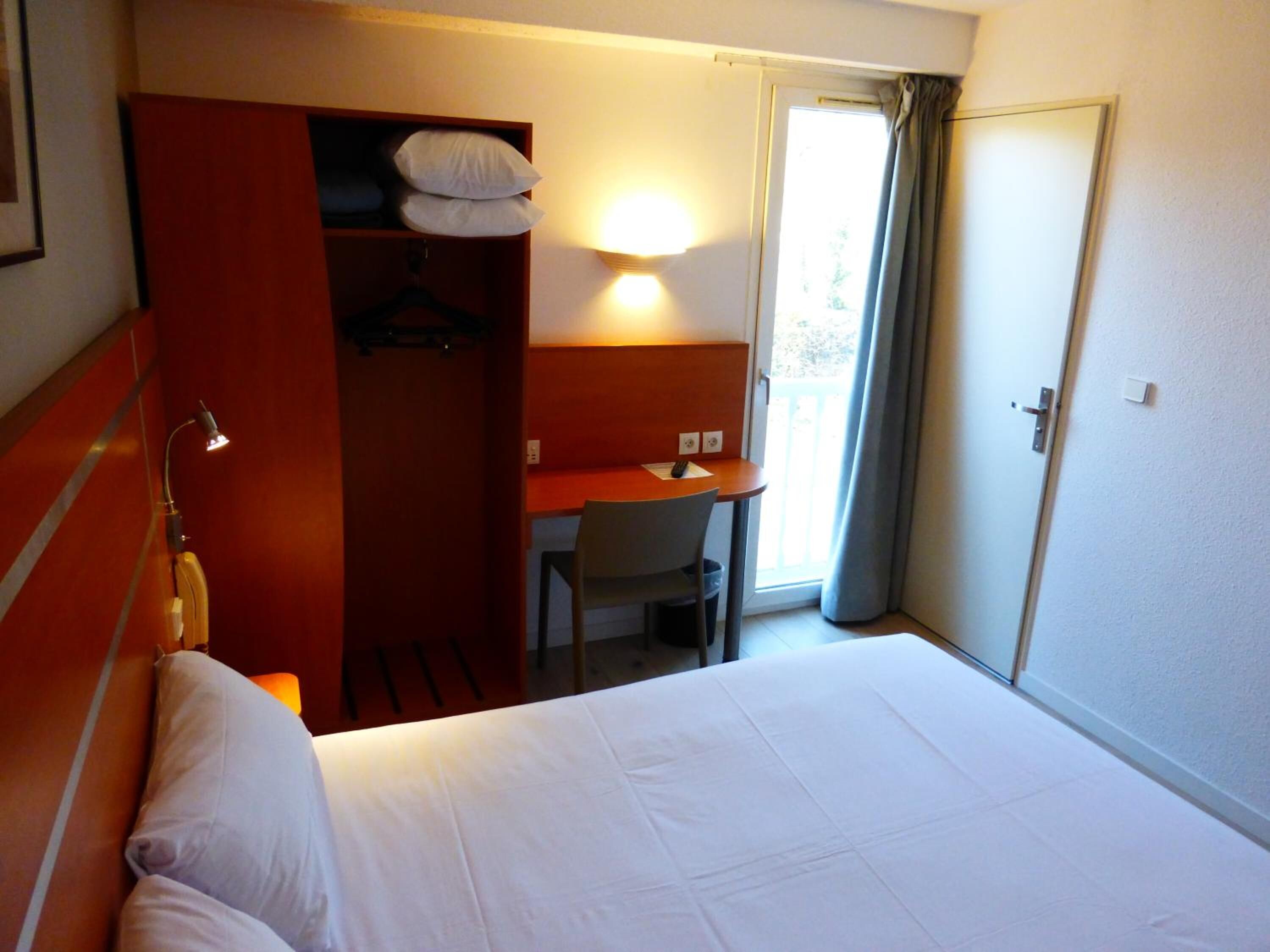 A hotel room with a bed, a desk with a chair, a wardrobe, and a window with curtains.