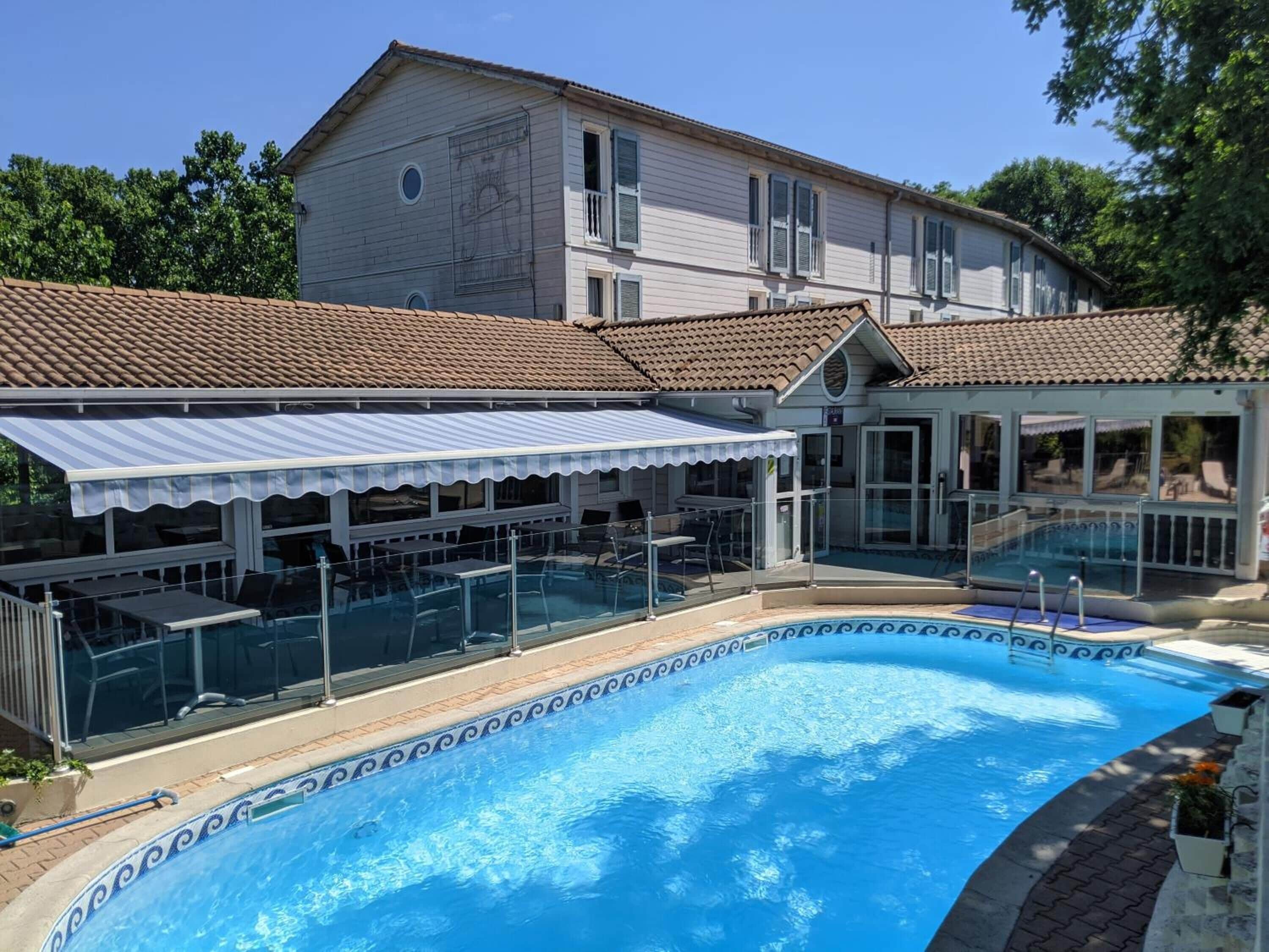 Piscina com deck ladrilhado, área de estar coberta e um edifício com telhado ladrilhado ao fundo.