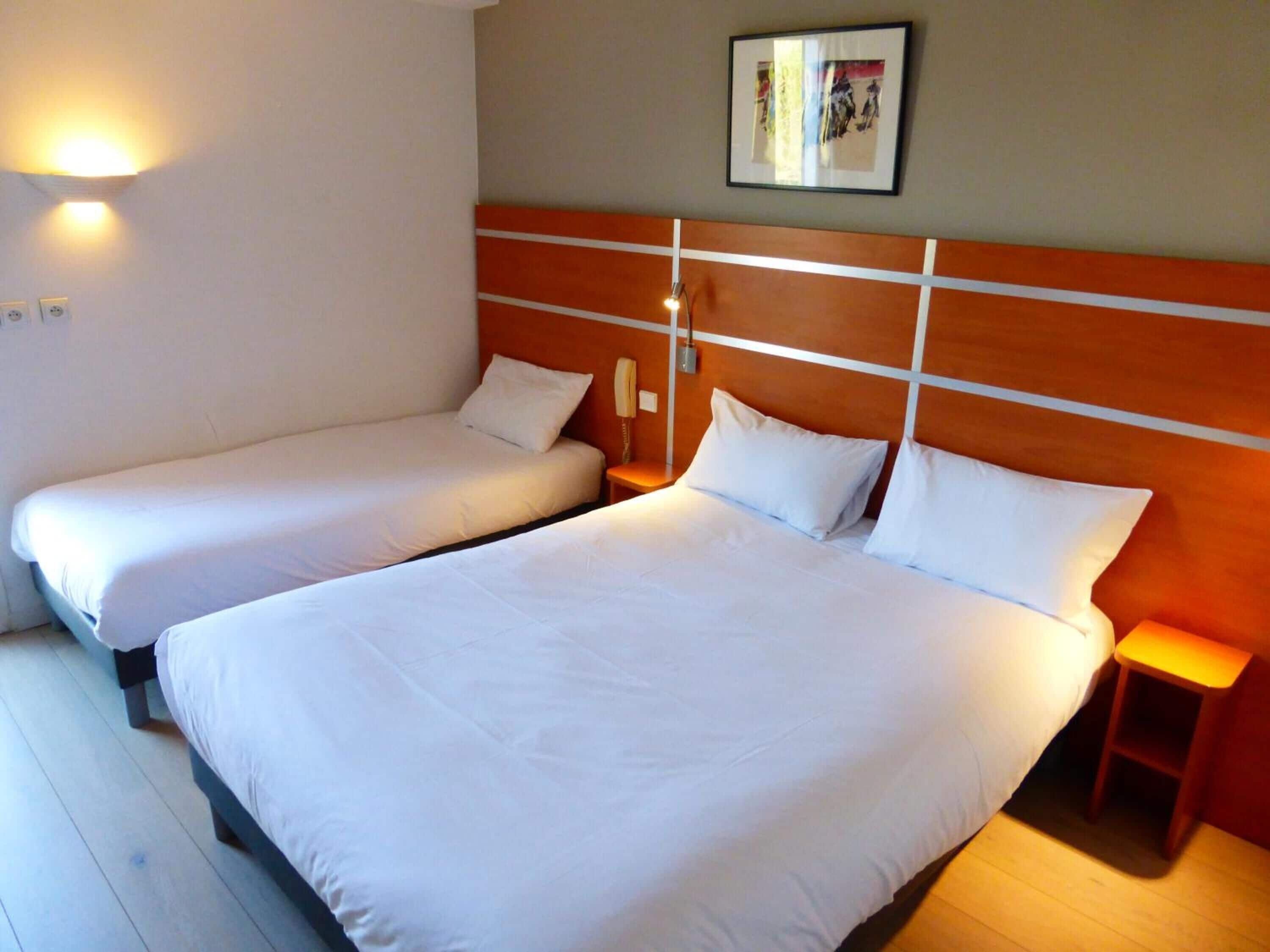 Une chambre d’hôtel avec deux lits simples, une tête de lit en bois, des tables de chevet et un luminaire fixé au mur.