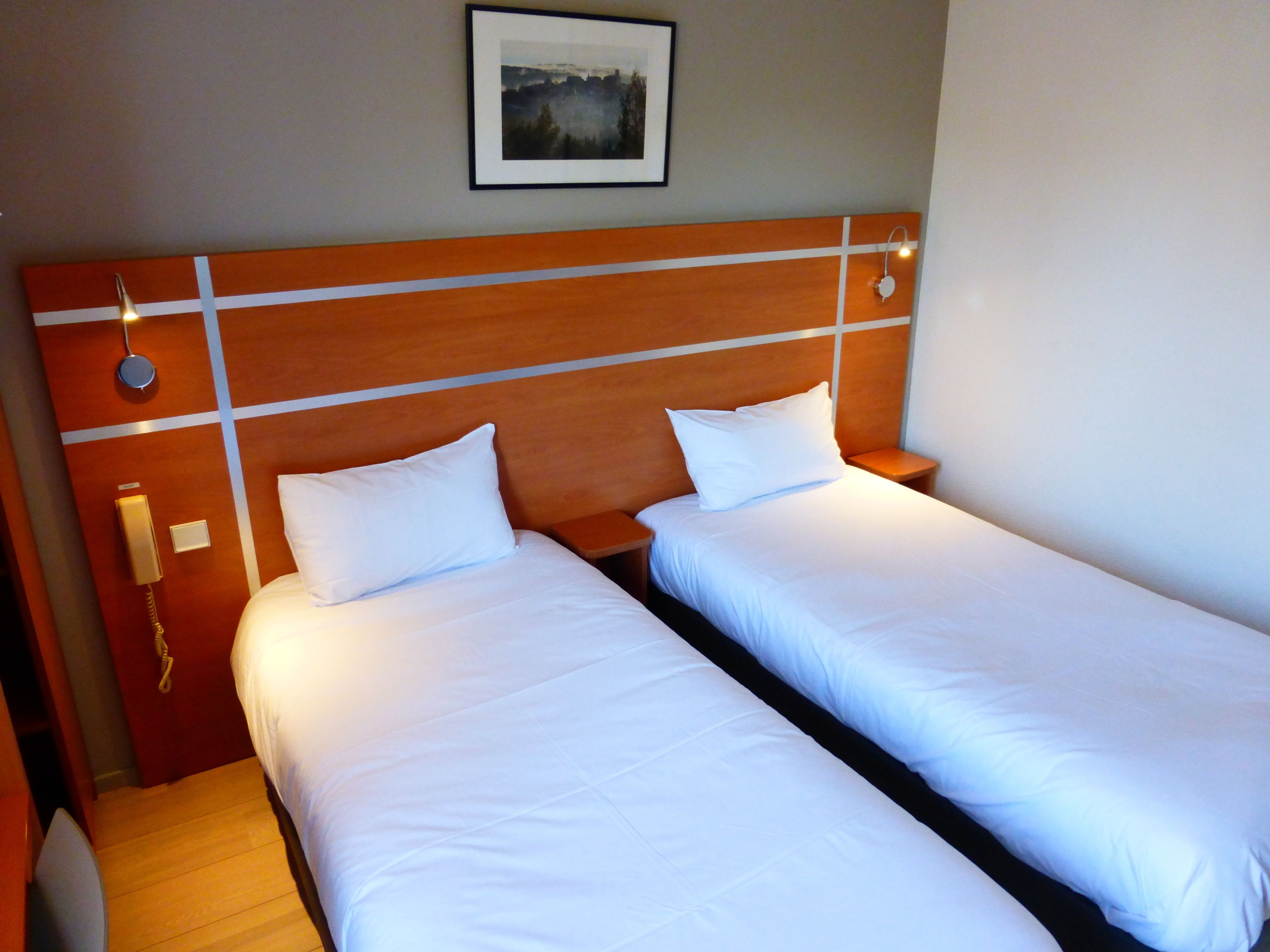 Une chambre d’hôtel avec deux lits simples, une tête de lit en bois, un téléphone sur la table de chevet et un tableau encadré au mur.