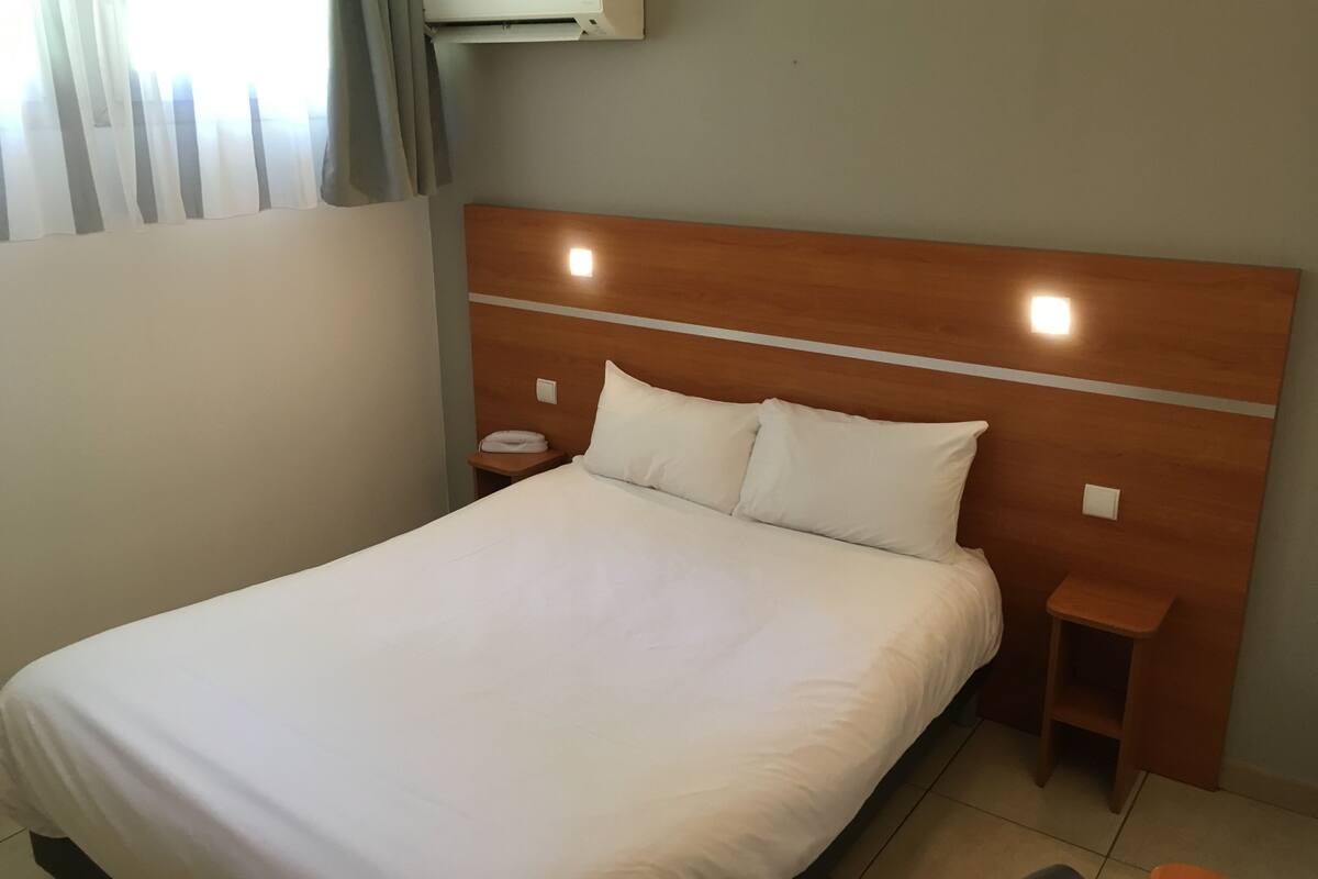 A hotel room with a wooden headboard, a white bed, a nightstand with a phone, and an air conditioning unit on the wall.