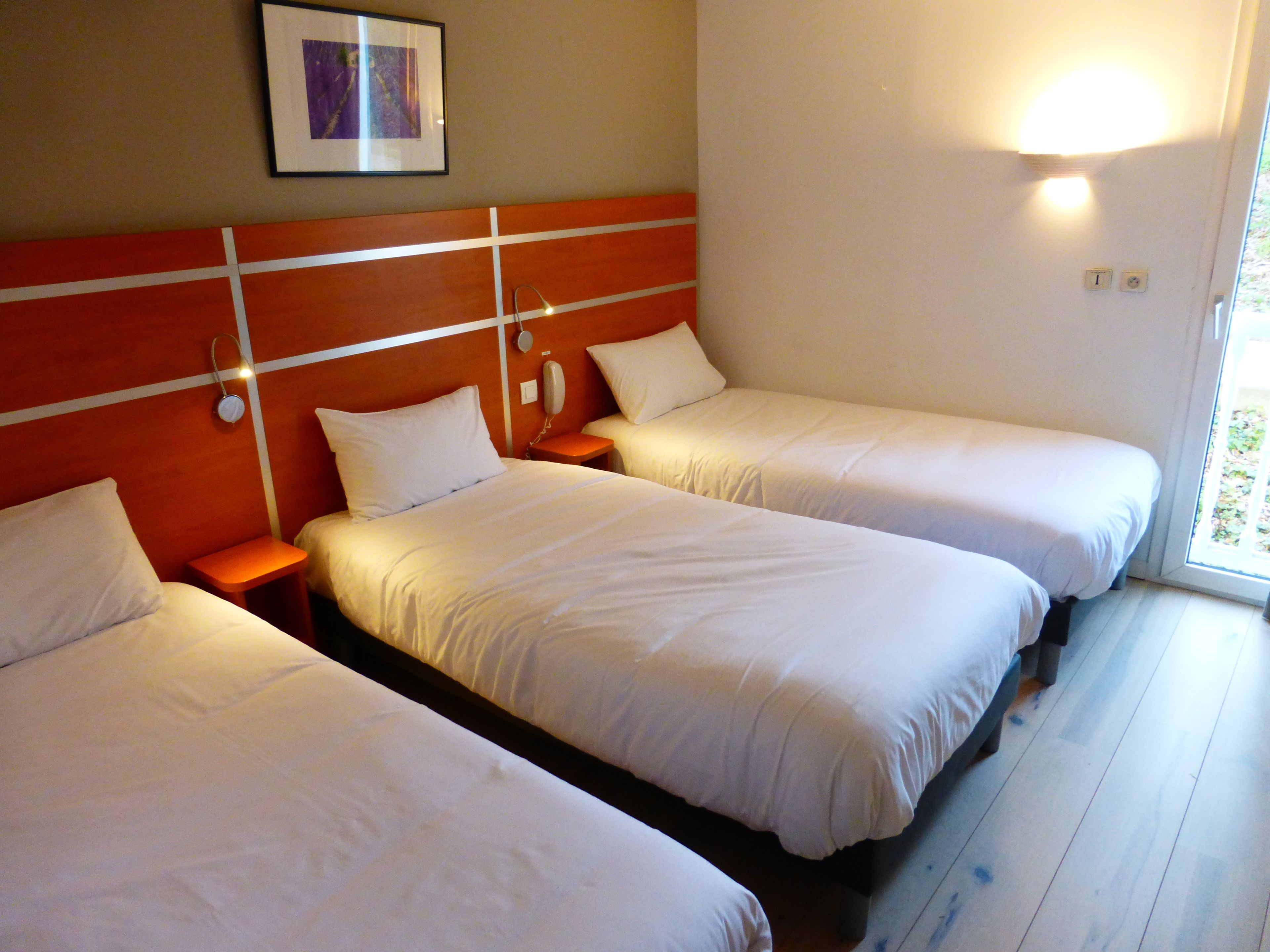 Ett hotellrum med tre enkelsängar, ett nattduksbord med en lampa, en inramad tavla på väggen och ett fönster med utsikt över grönområden.