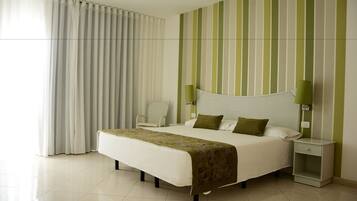 A hotel room with a large bed, a nightstand, a chair, and a view of the outside through sheer curtains.