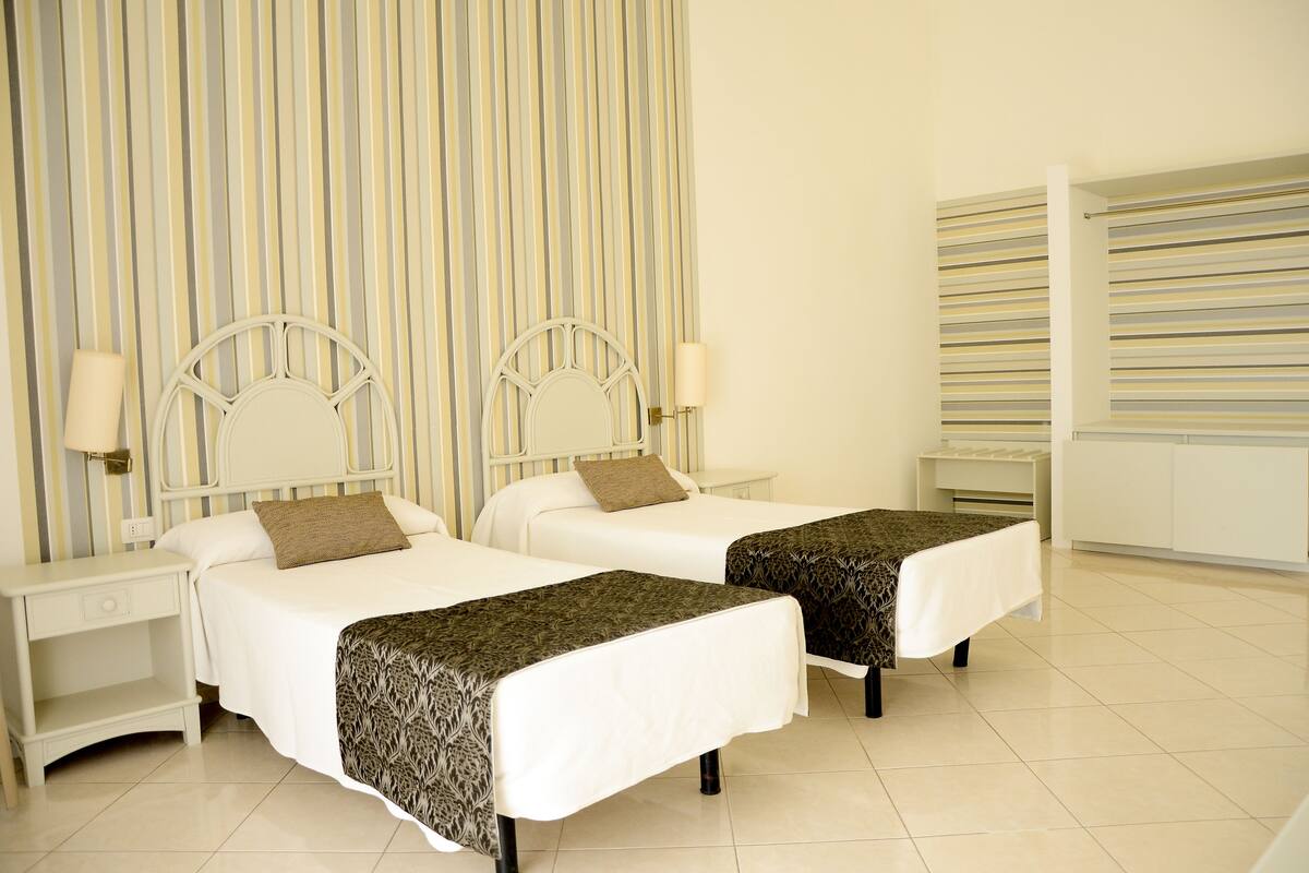 Quarto de hotel com duas camas de solteiro, uma mesa de cabeceira, uma luminária e cortina listrada.