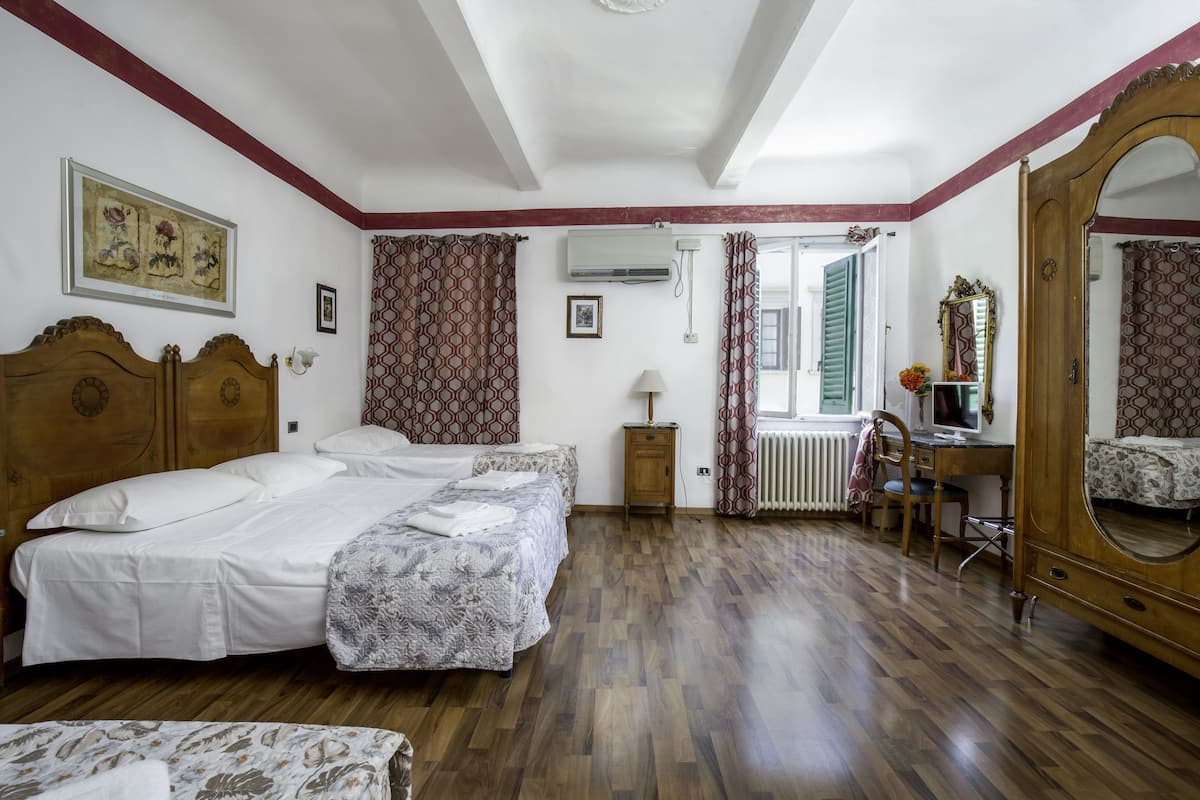 A hotel room with two beds, a wooden floor, a desk with a computer, a mirror, and a window with curtains.