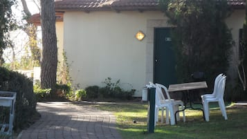 Een verhard pad dat leidt naar een gebouw met een betegeld dak en een groene deur. Buiten staan witte plastic stoelen en een tafel.