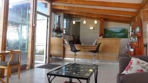 Un área de recepción con techo de madera, un mostrador de recepción, dos sillas negras, una mesa de vidrio y una alfombra roja.