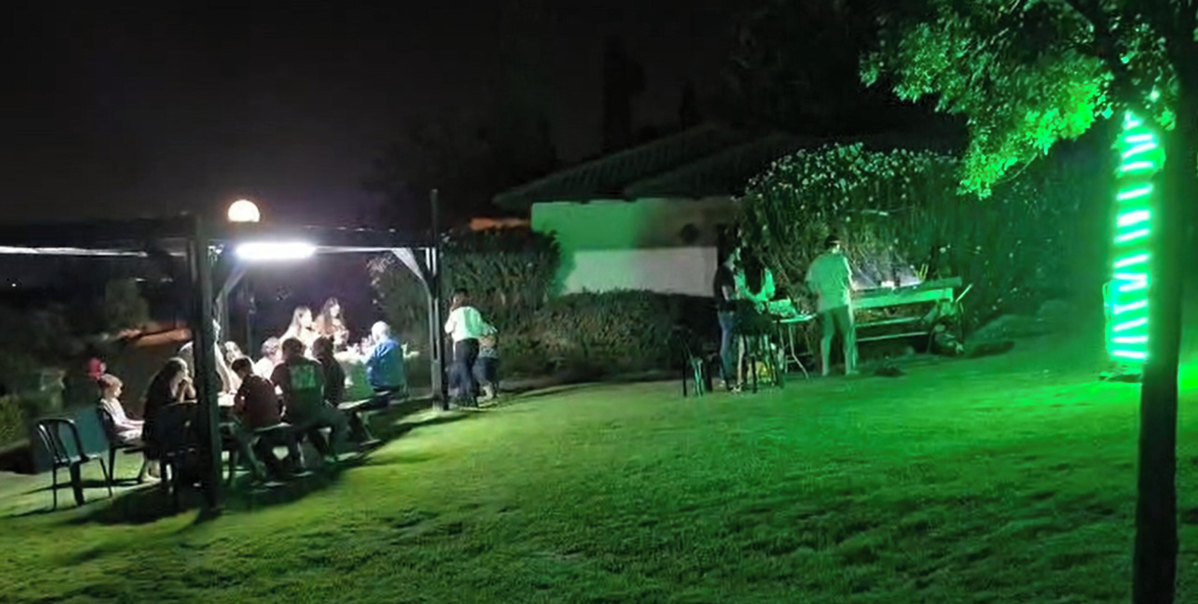 An outdoor area with a covered seating area and a table, surrounded by greenery and a hedge.