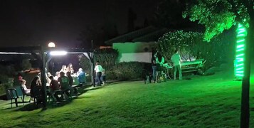 Espacio exterior con zona de estar cubierta y una mesa, rodeado de follaje y un seto.