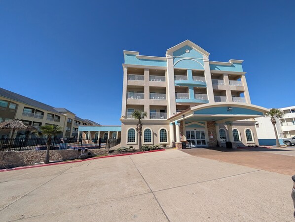 Un hotel de varios pisos con entrada central, balcones y zona de piscina.