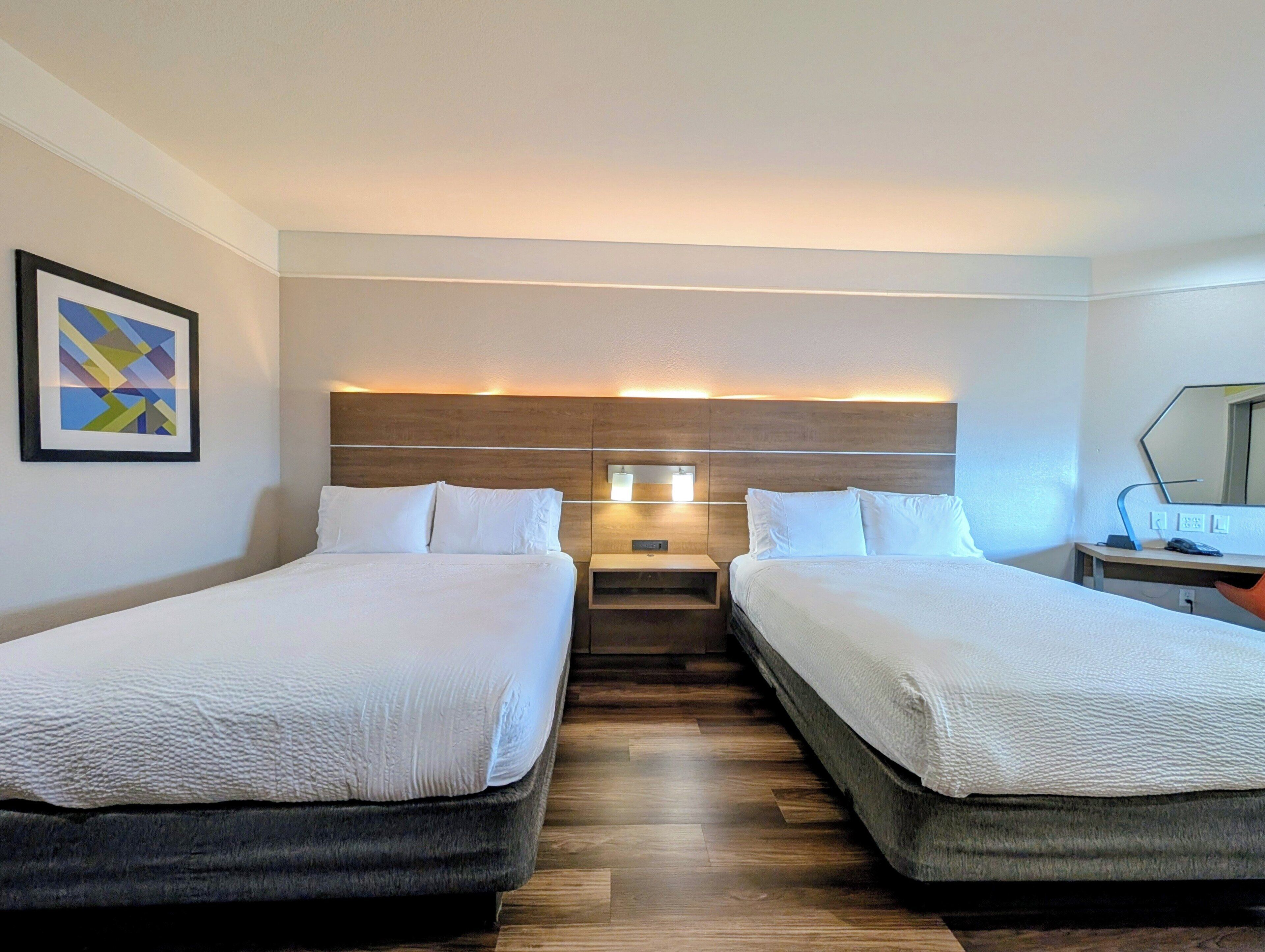 A hotel room with two beds, a wooden headboard, a bedside table with a lamp, and a framed picture on the wall.