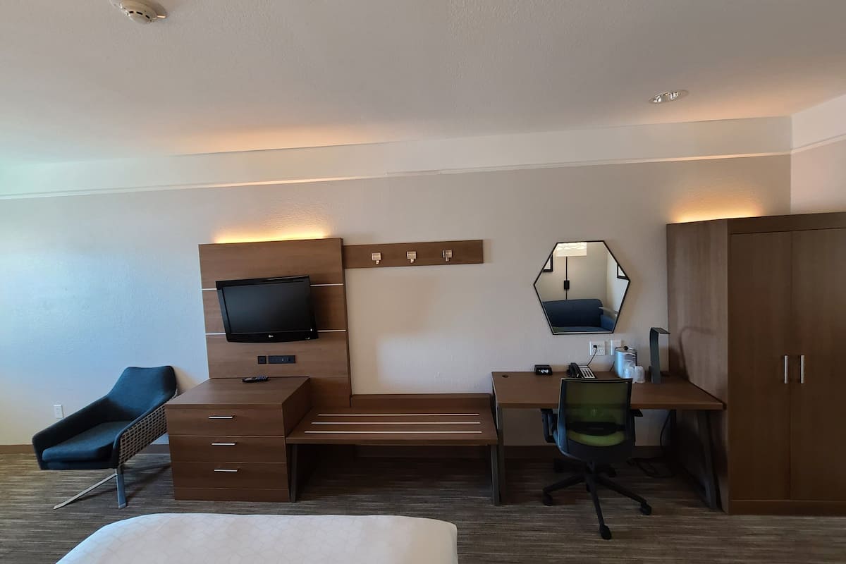 A hotel room with a bed, a desk with a computer, a chair, a TV, a mirror, and a wardrobe.
